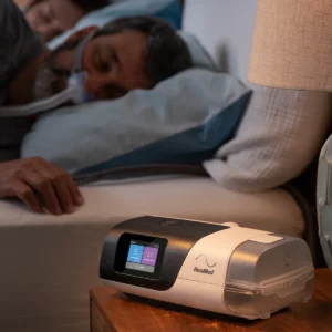 resmed-airsense-11-cpap ResMed AirSense 11 CPAP machine on bedside table while user sleeps with mask, illustrating sleep apnea therapy and PAP treatment at home.